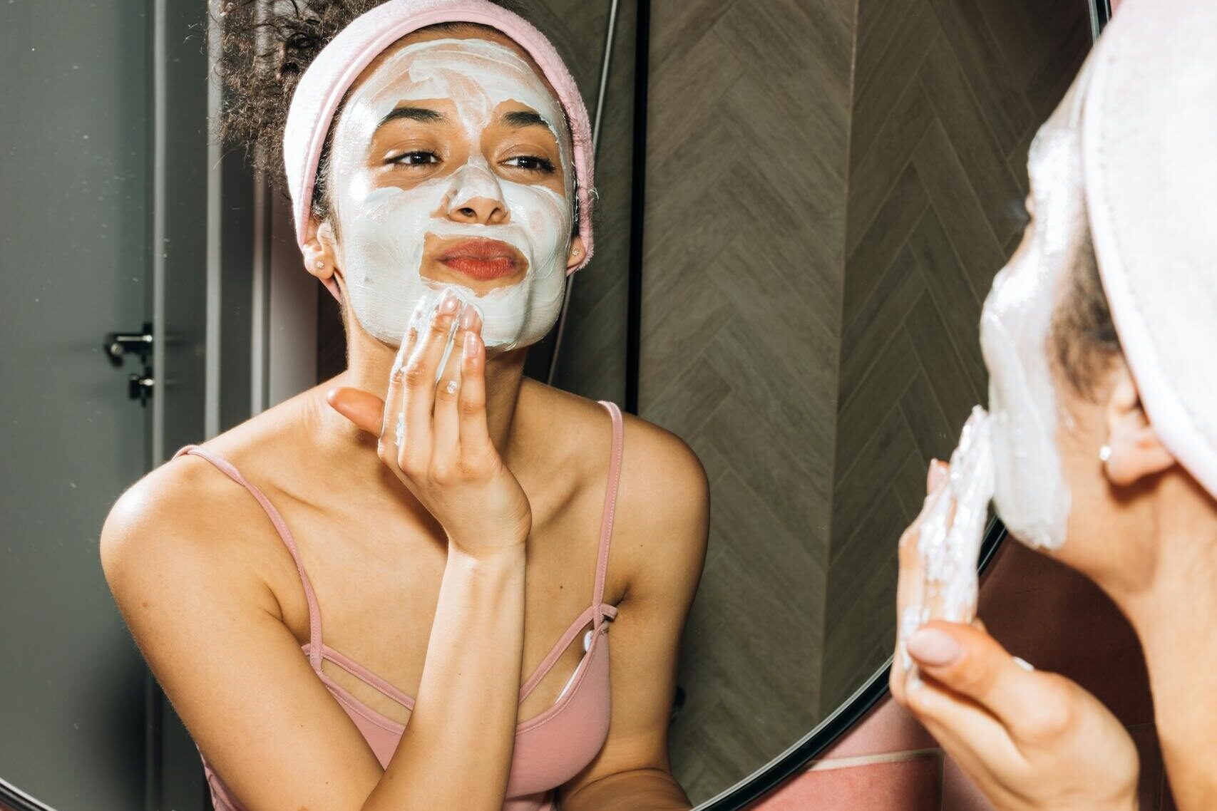 A young woman smiling while applying a facial mask during her home skincare routine.